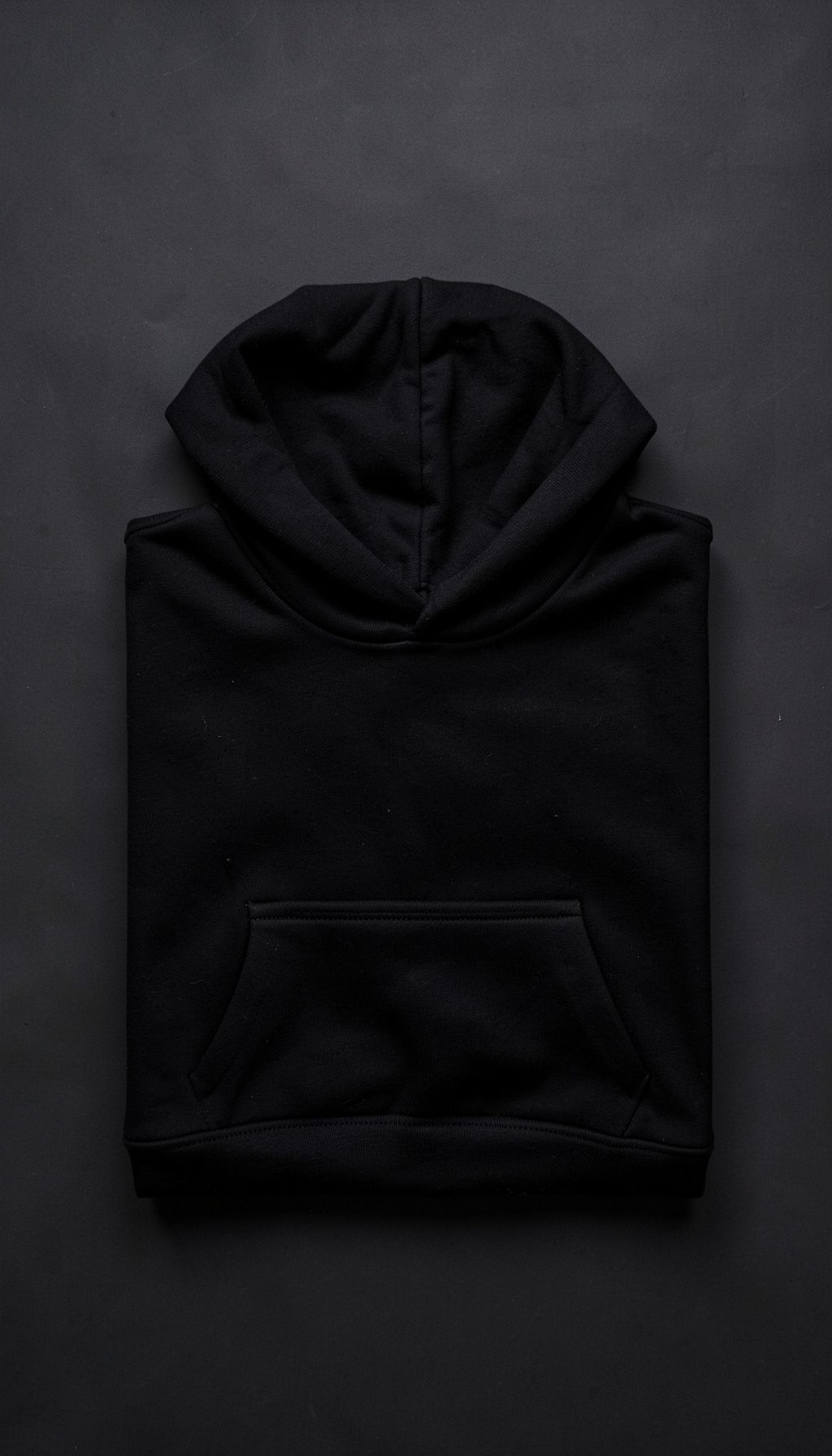 Overhead flat-lay of a black hoodie folded neatly on a dark surface, the fabric texture visible under soft directional light